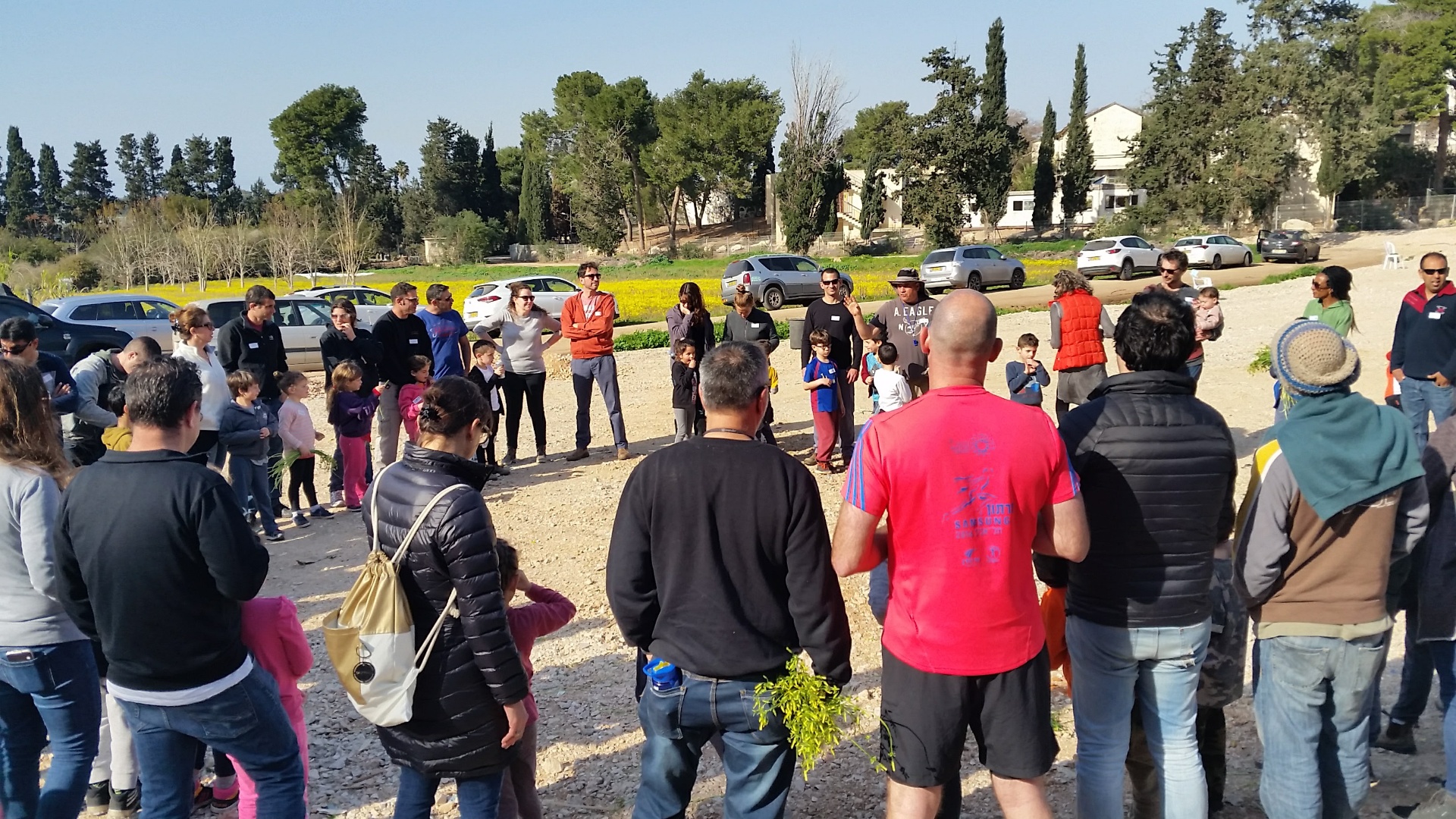 מפגש קהילתי בחוץ, משפחות וילדים מקשיבים למרצה בשדה פתוח
